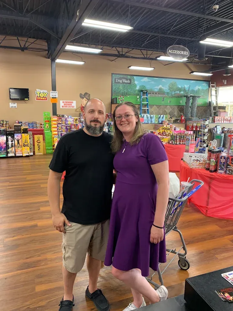 Two people posing together in a brightly lit store aisle, one in black and one in a purple dress.
