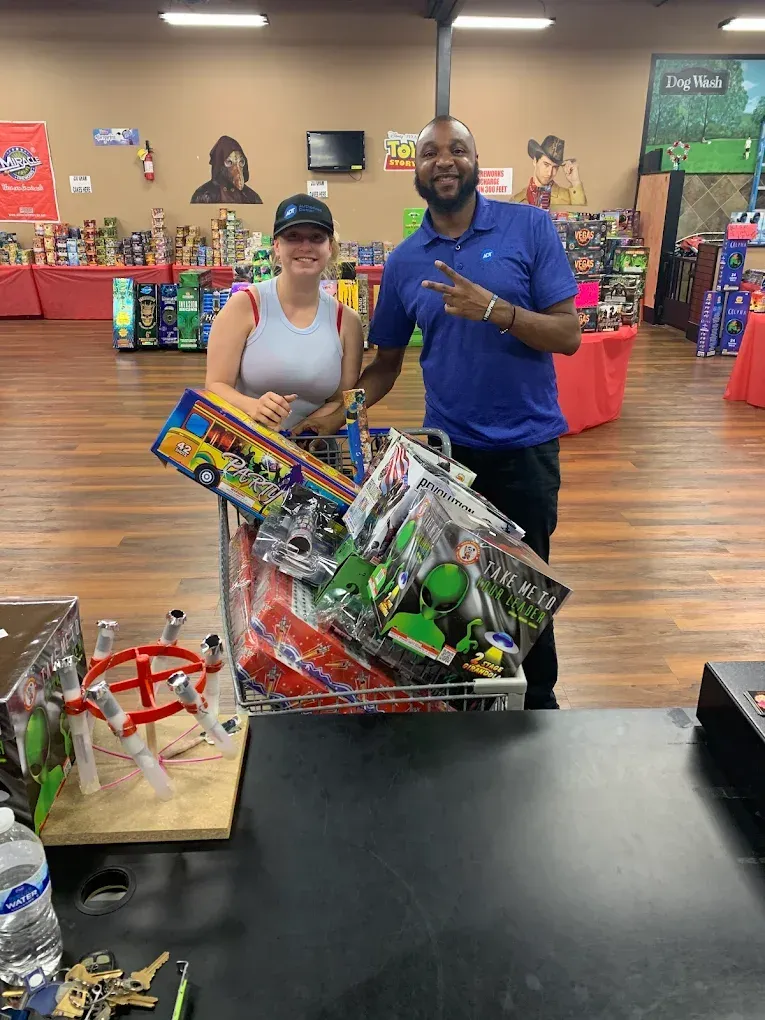 Two people stand in a store holding a large toy-filled gift basket in front of stacked gift displays.