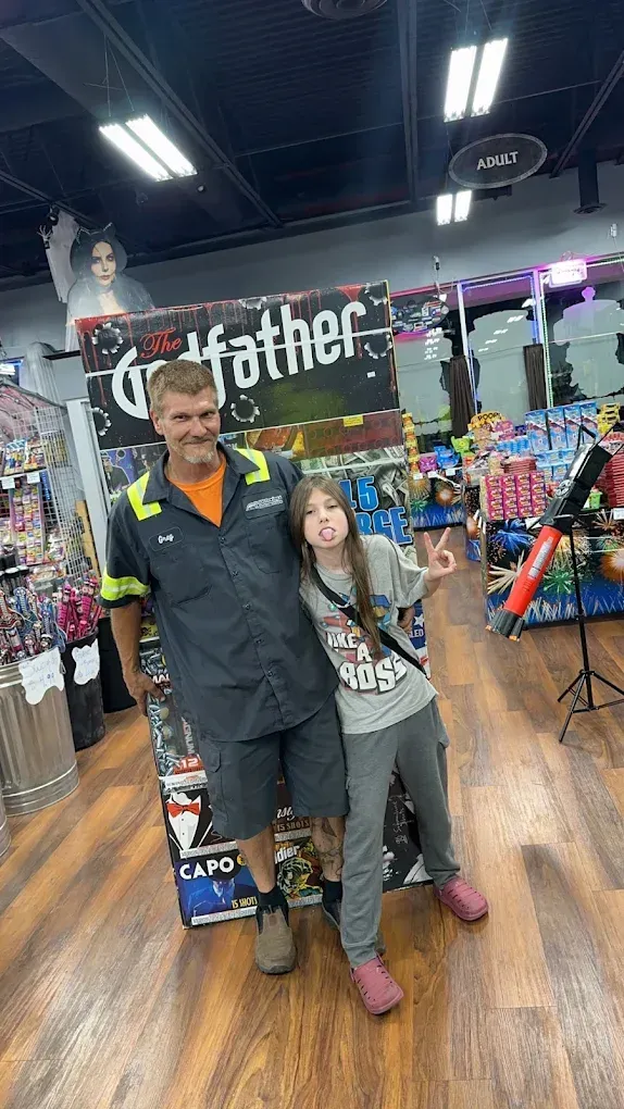 Man and child posing in a store aisle, with colorful shelves and a black “godfather” sign behind them.