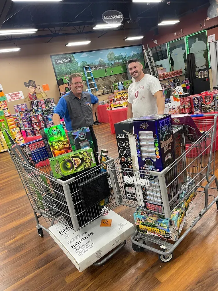 Two men pose with two full shopping carts in a toy store aisle.