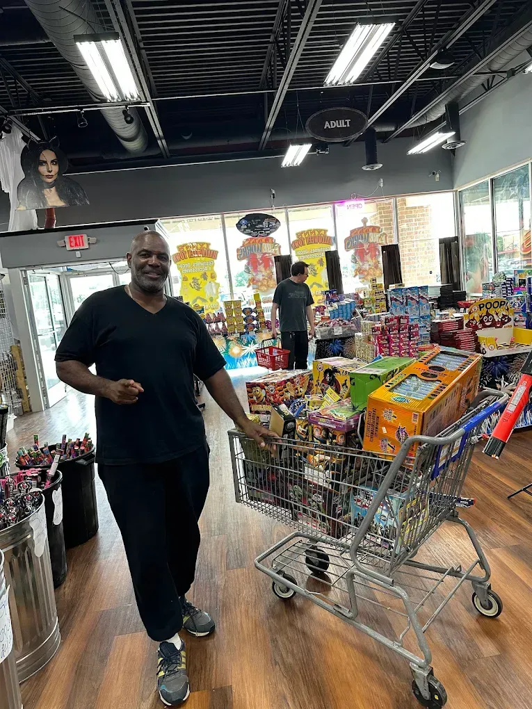 Man standing in a brightly lit store aisle beside shopping carts filled with colorful items.