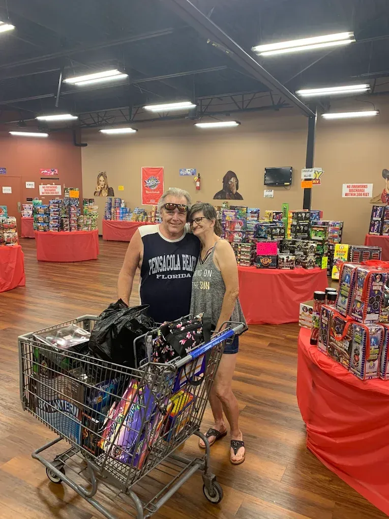 Two people posing in a toy store aisle beside a shopping cart full of colorful merchandise.