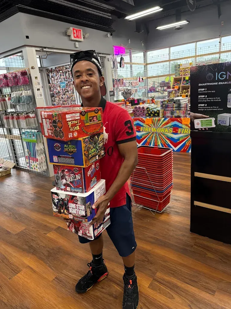 Smiling man in a toy store, holding a tall stack of colorful boxed toys and standing on a wooden floor.