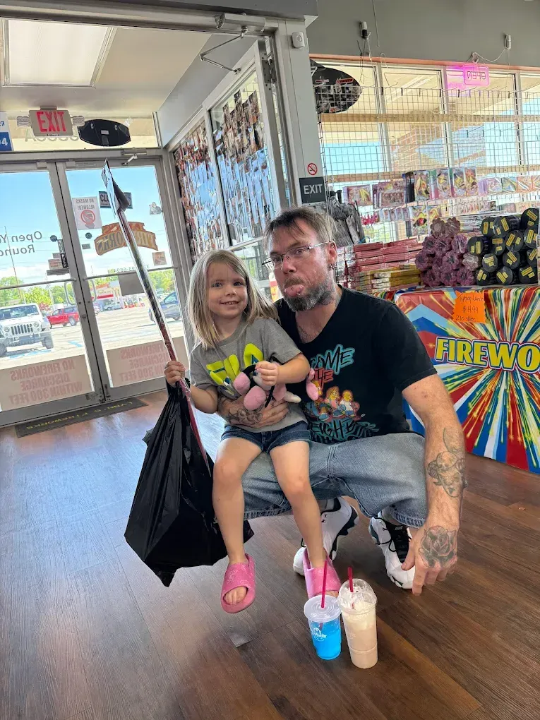 Two children sitting on a bench in a store, one holding colorful cups with a bright prize wheel behind them.