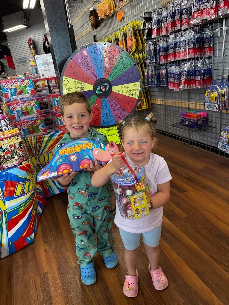 Two children smiling in a colorful store aisle, holding rainbow candy and standing by a prize wheel.