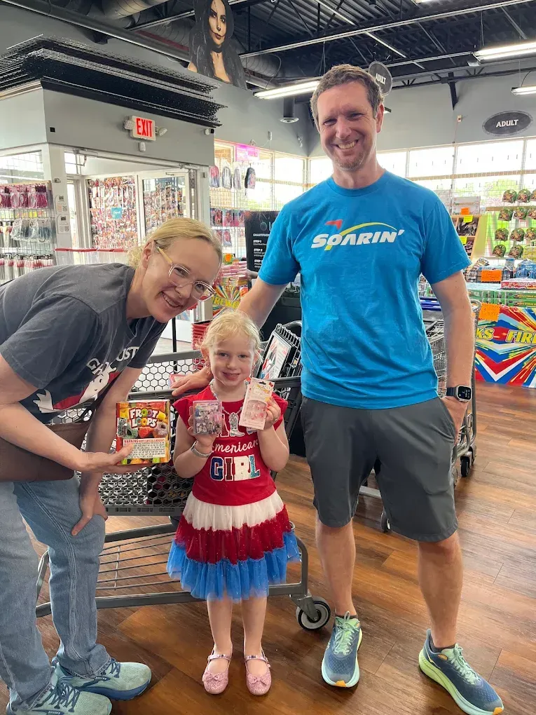 Three people posing in a store aisle, with the center person holding a small cup and wearing a red-white-blue dress.