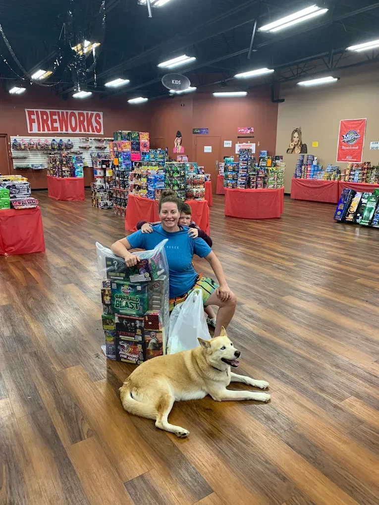 Person kneeling with a dog in a fireworks store aisle, surrounded by colorful fireworks displays.
