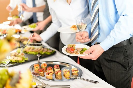 tavola con buffet con del cibo, in piedi un uomo con un bicchiere di vino in mano e un piatto e dietro altre persone che si servono