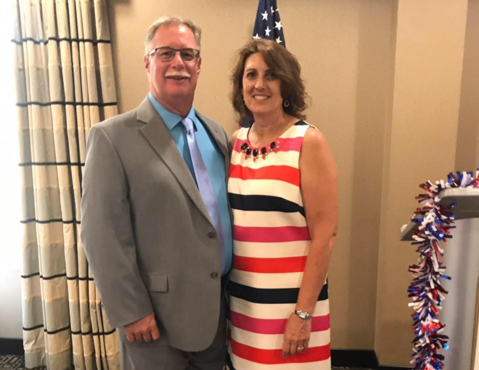 John Roth and his wife are posing for a picture in front of an american flag.