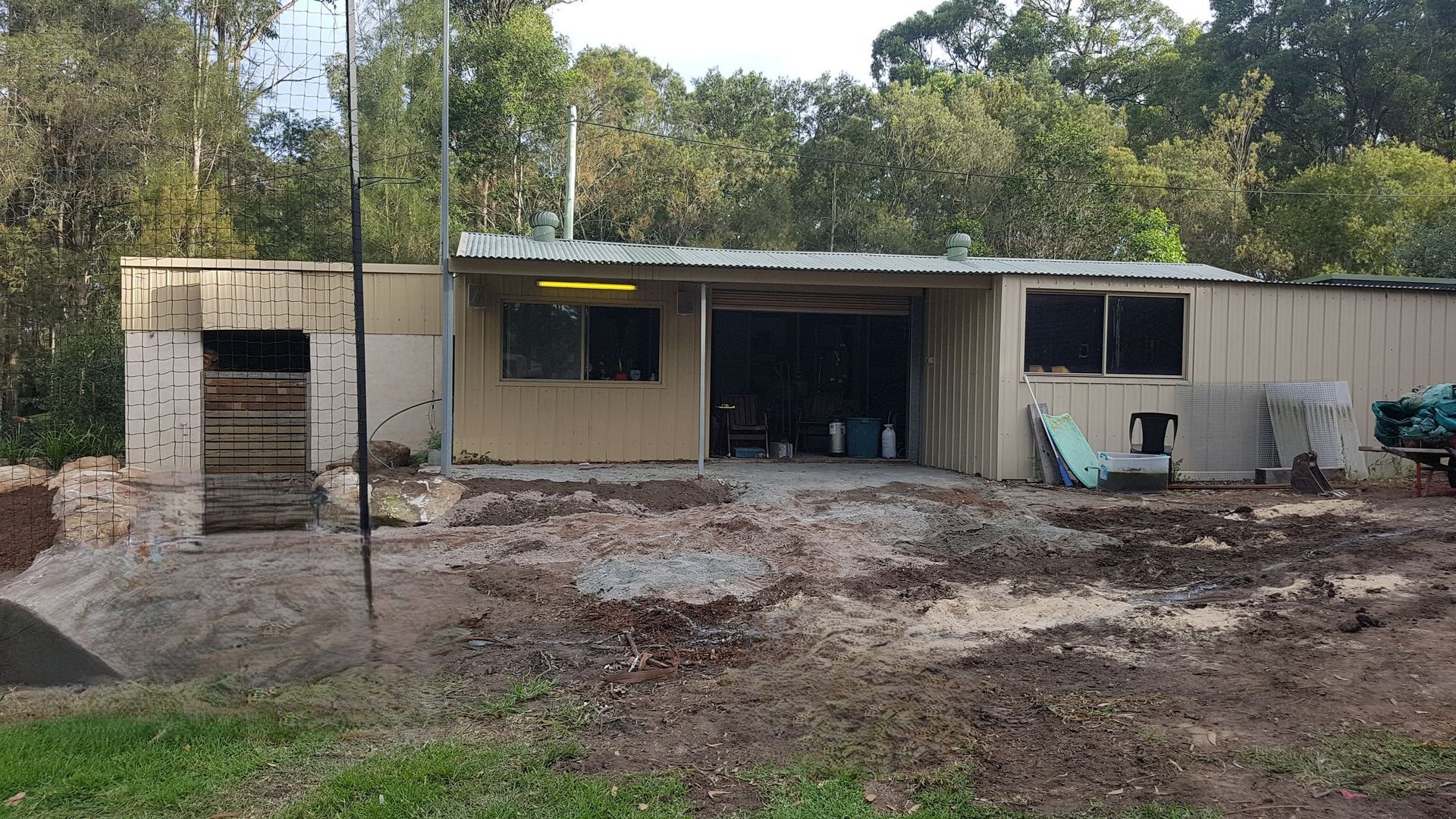A weathered, beige-colored building with a carport and unfinished area; surrounded by trees and dirt.