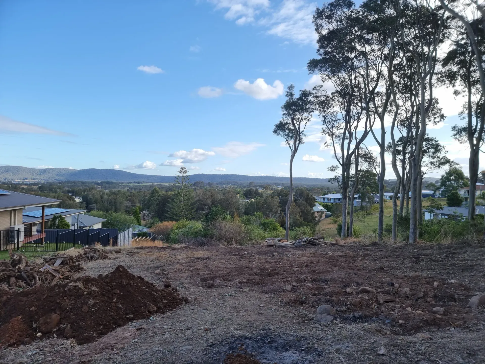 Cleared land with a scenic view, trees, and houses under a partly cloudy sky.