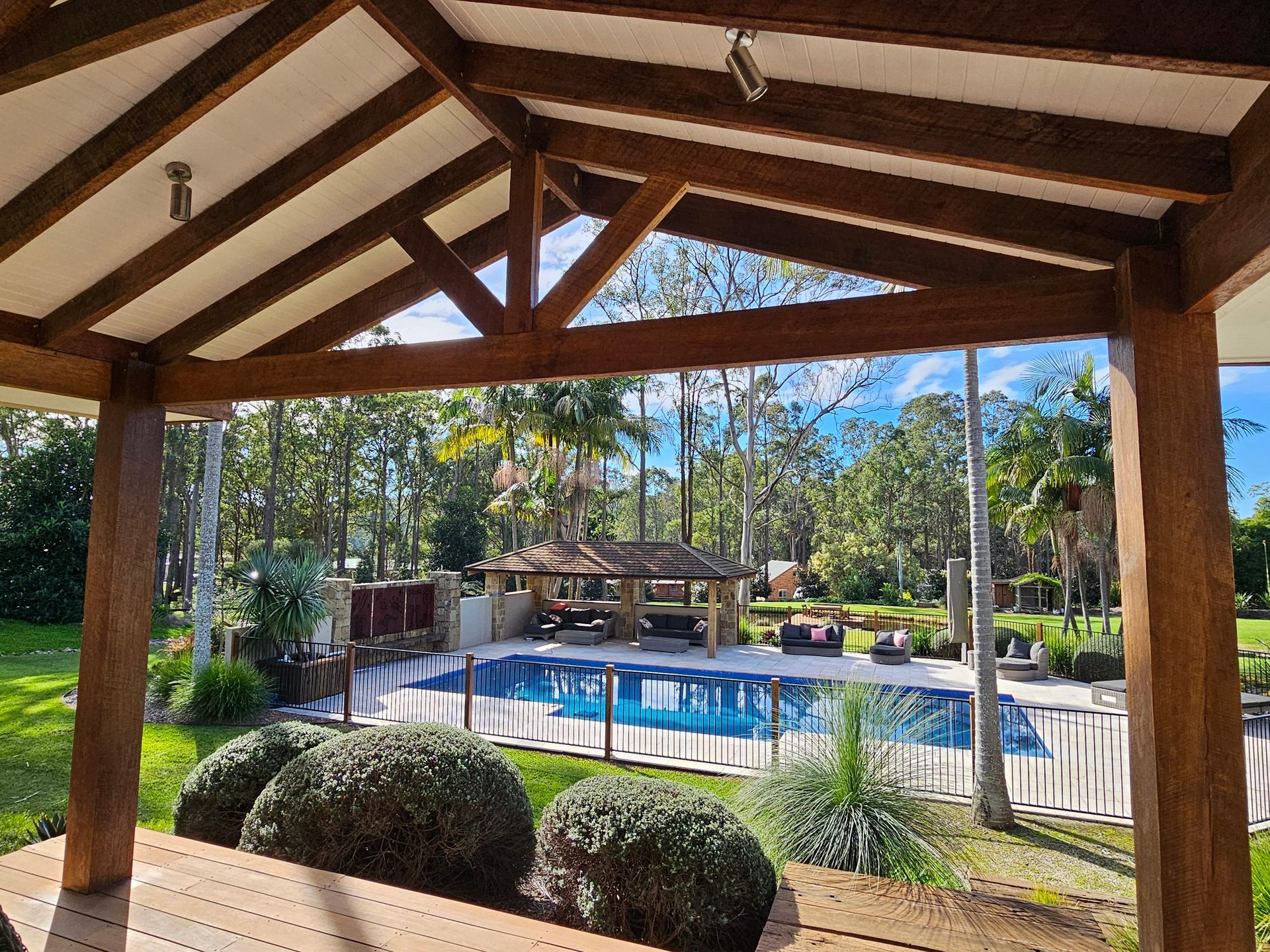 Wooden pergola overlooking a pool, with trees and a sunny sky in the background.