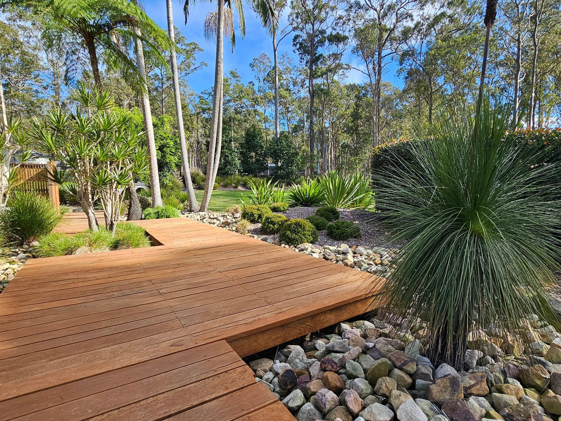 Wooden walkway through a lush garden with trees, plants, and rocks under a blue sky.