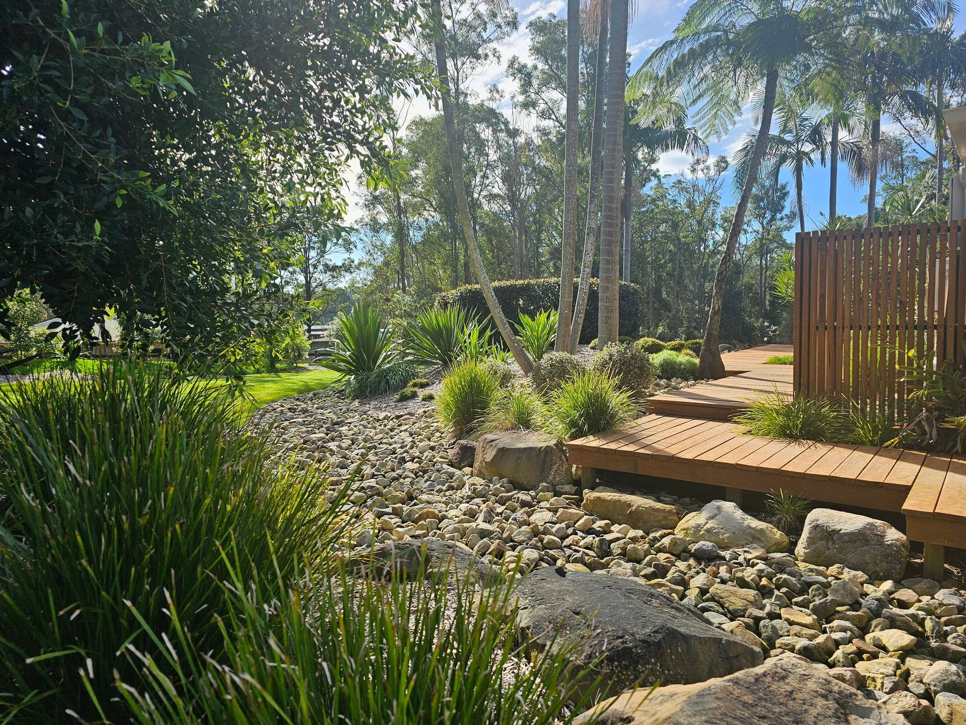 Lush garden with dry creek bed, wooden walkway, and various green plants and trees under a bright sky.
