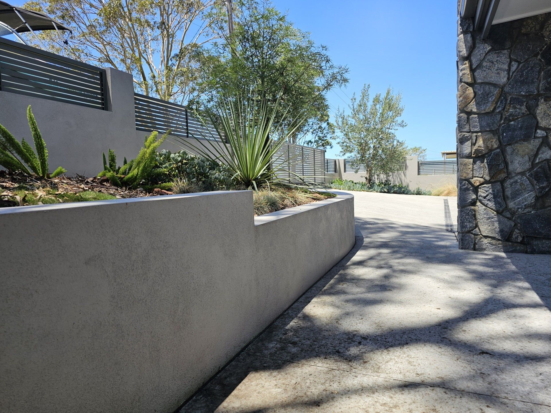 Concrete wall and path in a landscaped outdoor setting with trees and blue sky.