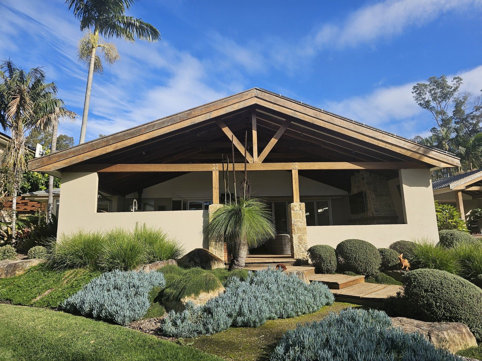 Modern two-story house with balcony, wooden accents, and stone landscaping. Greenery and large rocks in the foreground.