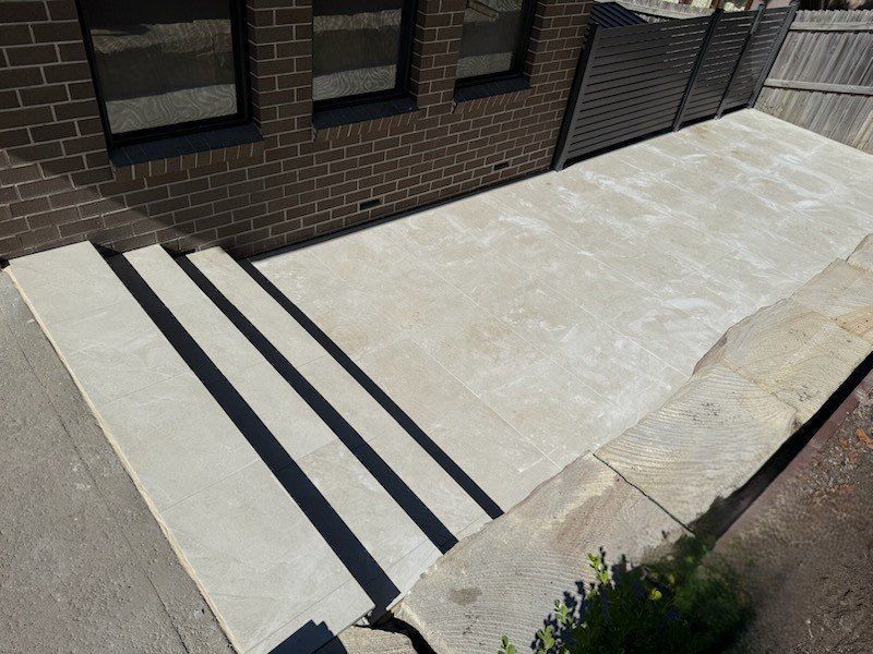 Concrete steps lead to a light-colored patio next to a brick wall with windows.
