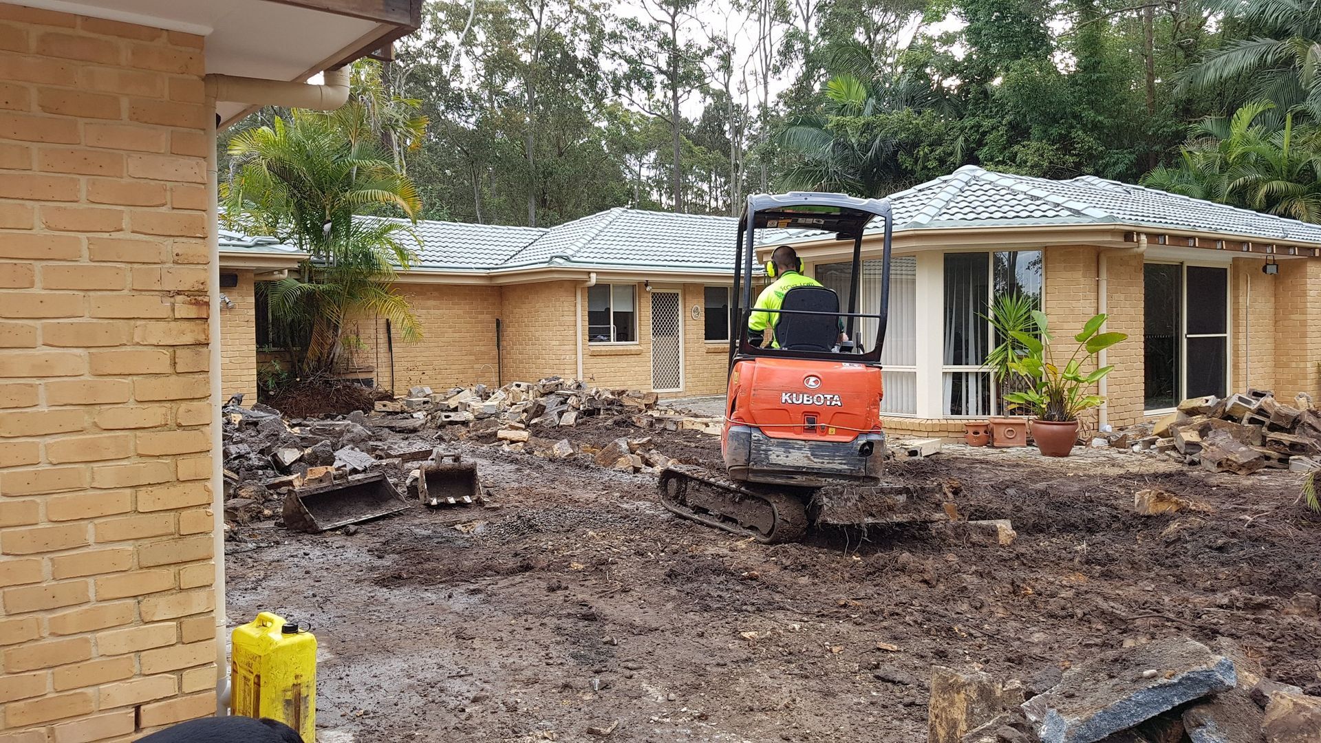 Mini excavator operating near a house, clearing debris. The worker wears a vest.