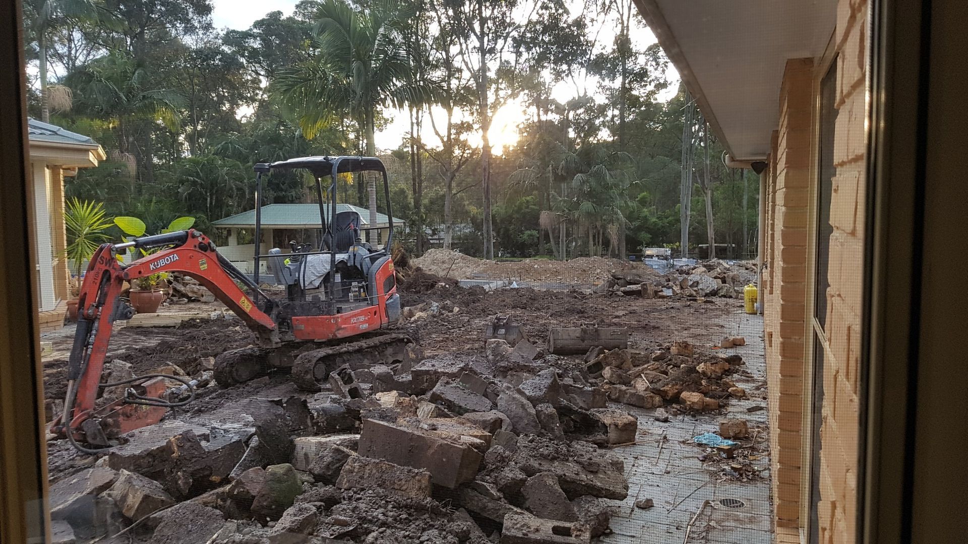 Mini excavator in a backyard, clearing debris, with the sun setting through trees.