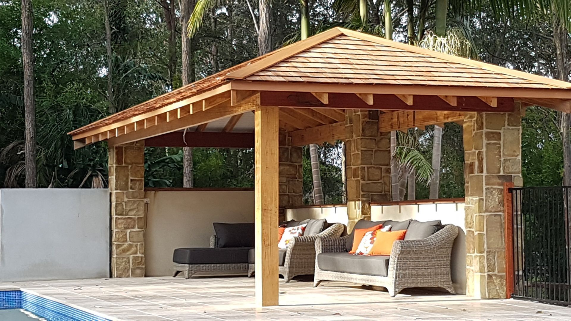 Gazebo with sandstone walls, timber frame, and shingled roof. Outdoor seating area with pool in foreground.