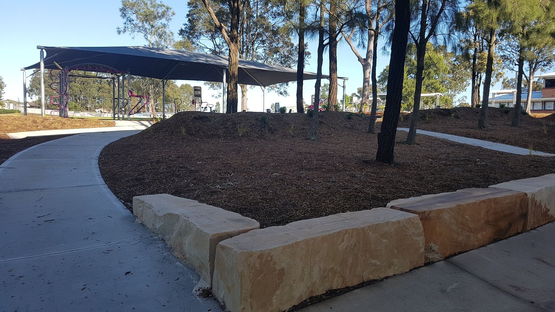 Concrete walkway curving around a raised garden bed with stone retaining wall. Trees and a covered shelter are in the background.