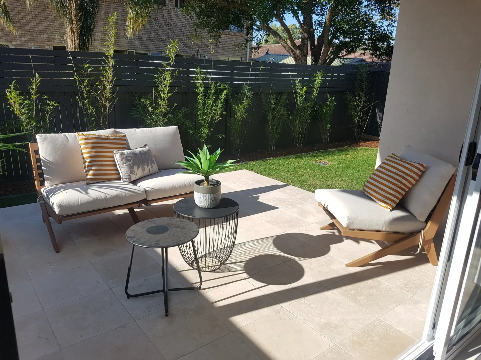 Patio with wood furniture and small tables, against a dark fence and green bushes.