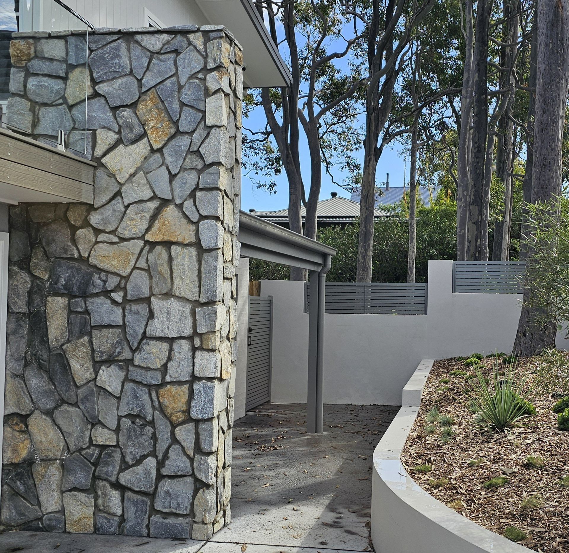 Stone wall facade next to a pathway leading to a carport and garden with trees.