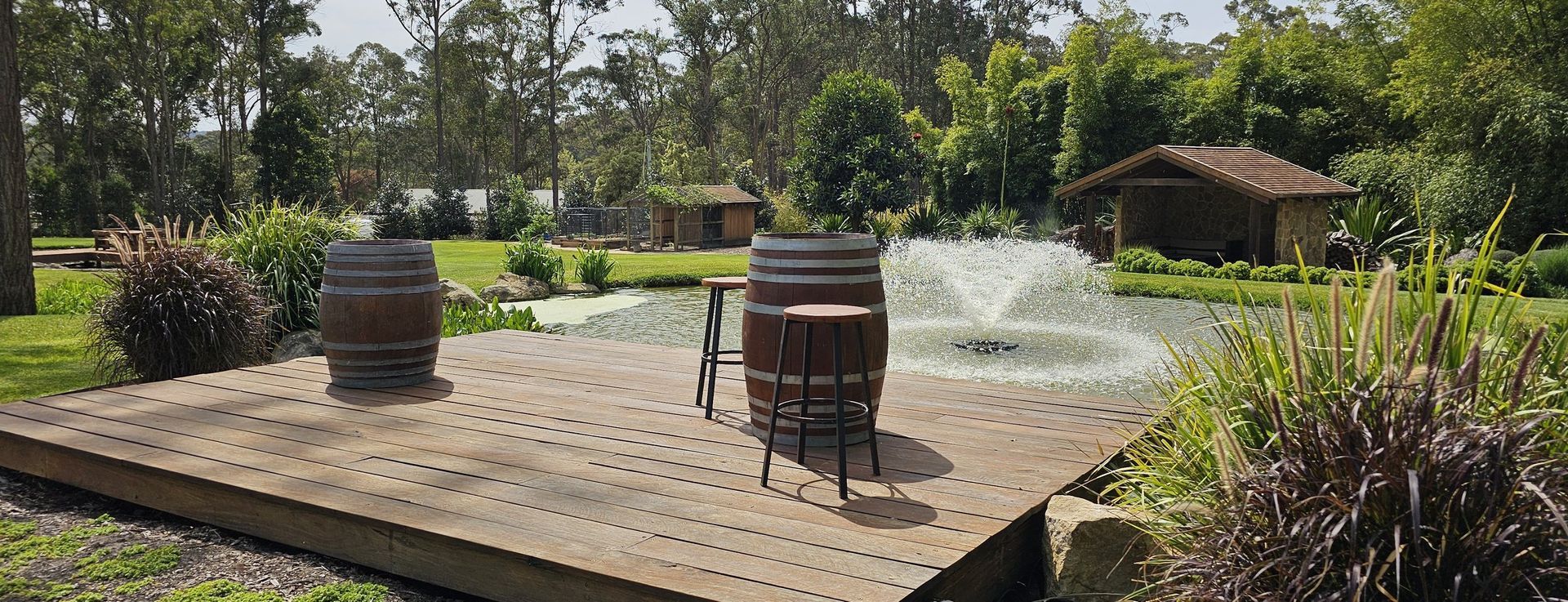 Wooden platform with two wine barrels, a table, and a fountain in a pond, surrounded by greenery.