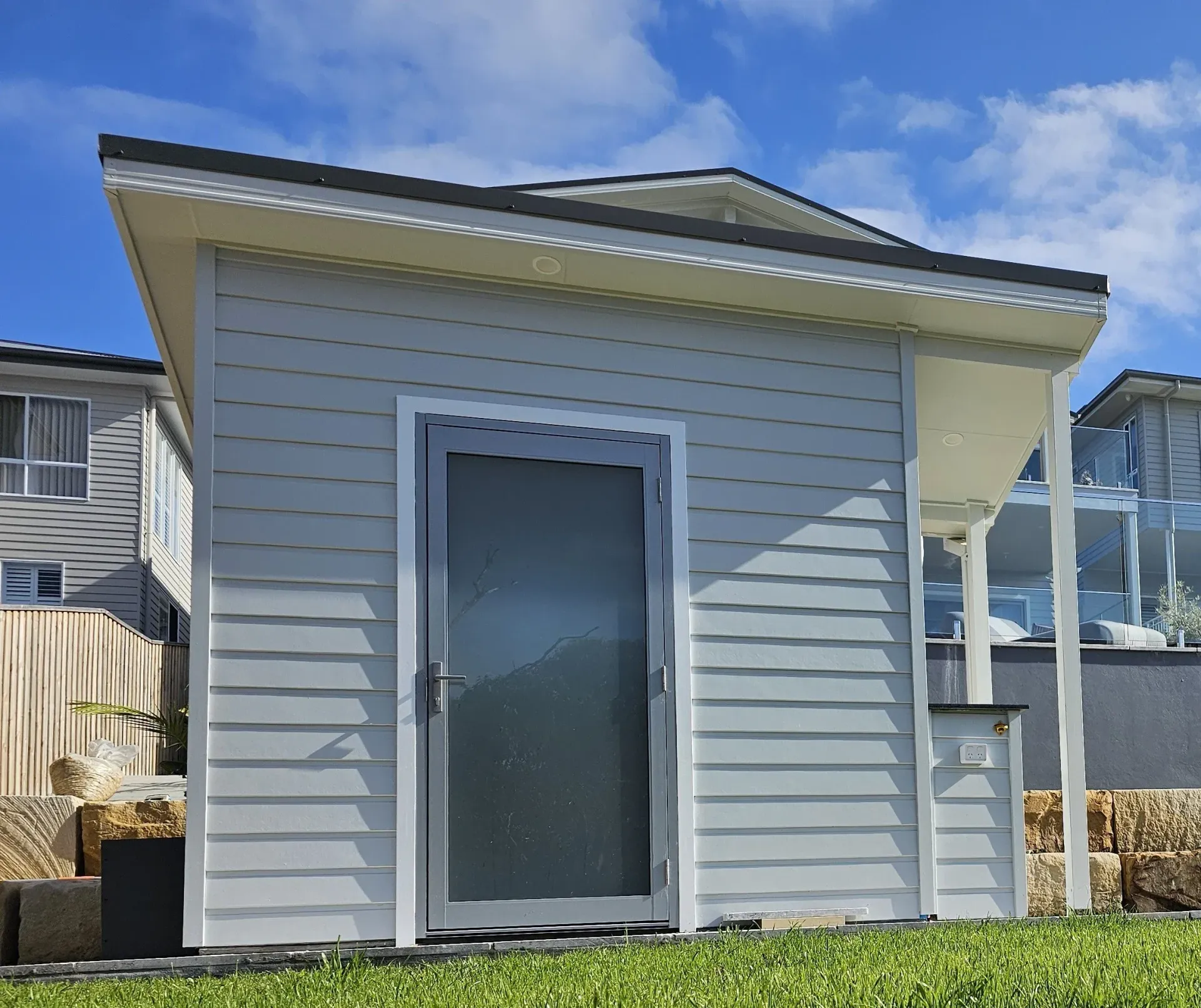 Small gray building with a door, facing a lawn, beside a larger building. Blue sky.