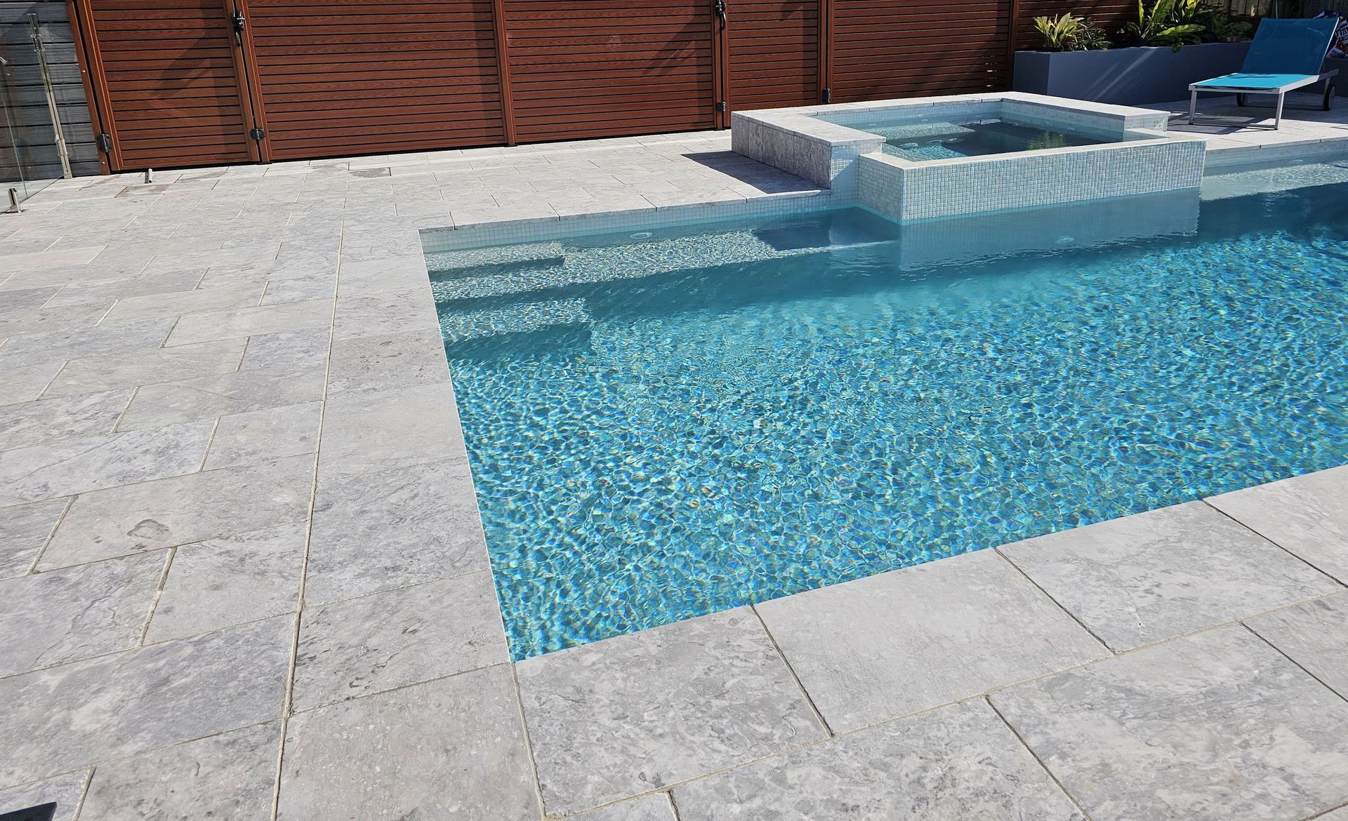 Swimming pool with light gray stone patio and raised spa, next to a brown wooden fence.