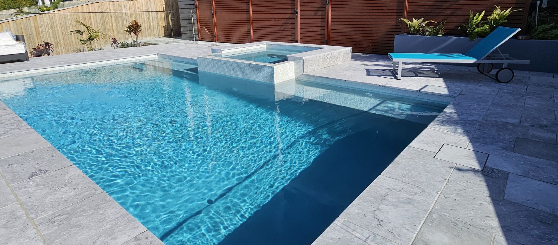 A turquoise pool and jacuzzi, surrounded by gray stonework and a lounge chair, outside on a sunny day.