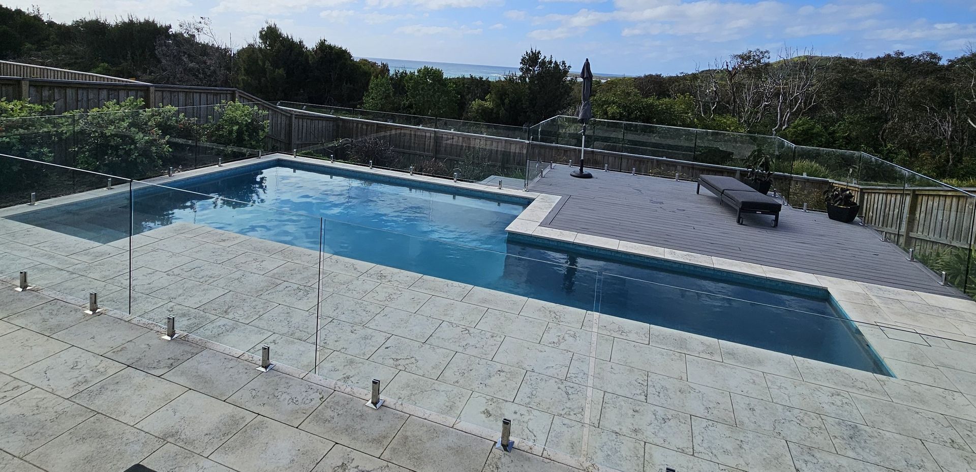 A rectangular pool surrounded by a patio and glass fencing, with trees and a view of the sea in the background.
