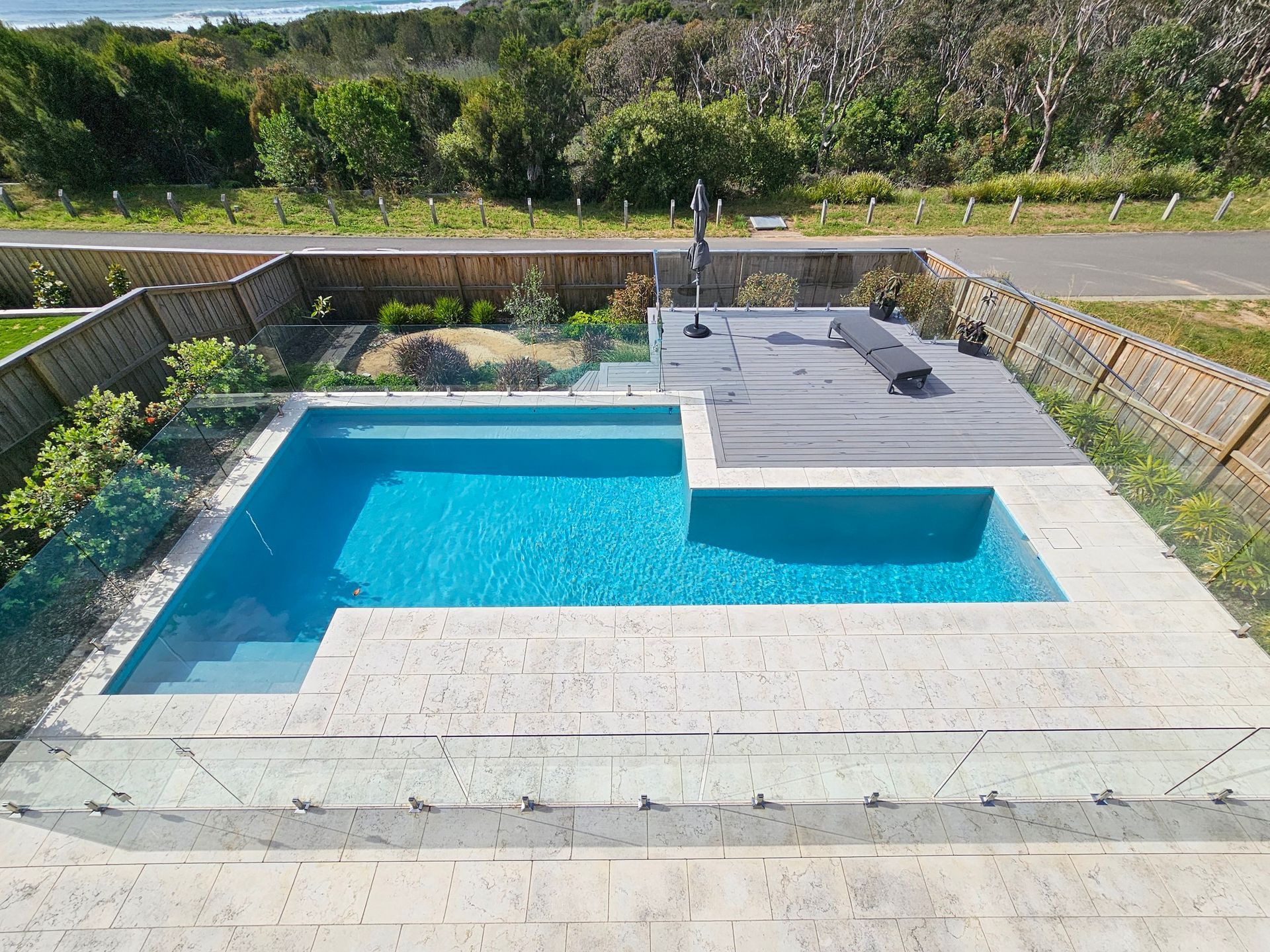 A swimming pool and spa with turquoise water and light stone surround, next to a backyard with greenery.