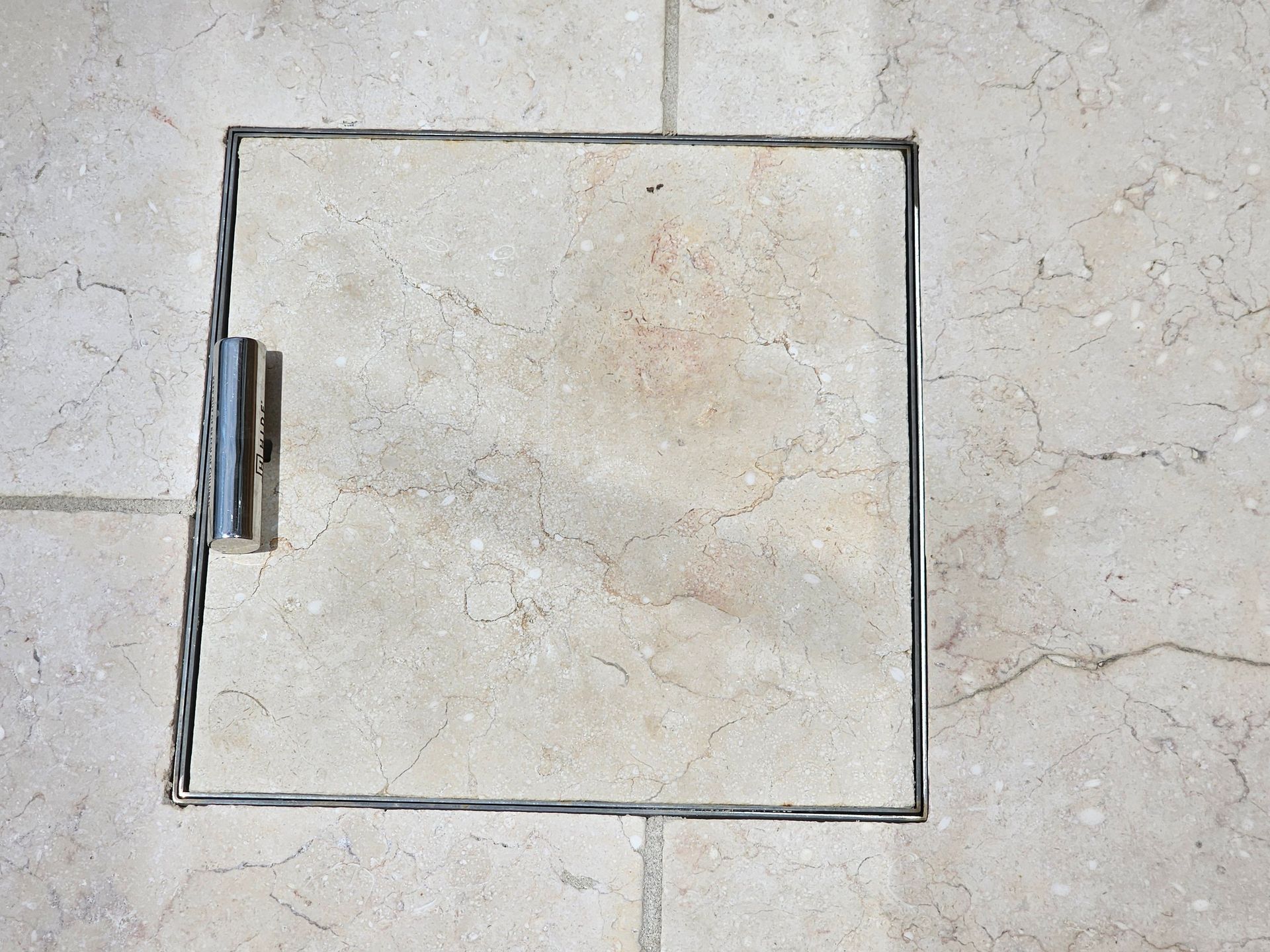 Square access panel on a tiled floor, with a metal handle and frame.