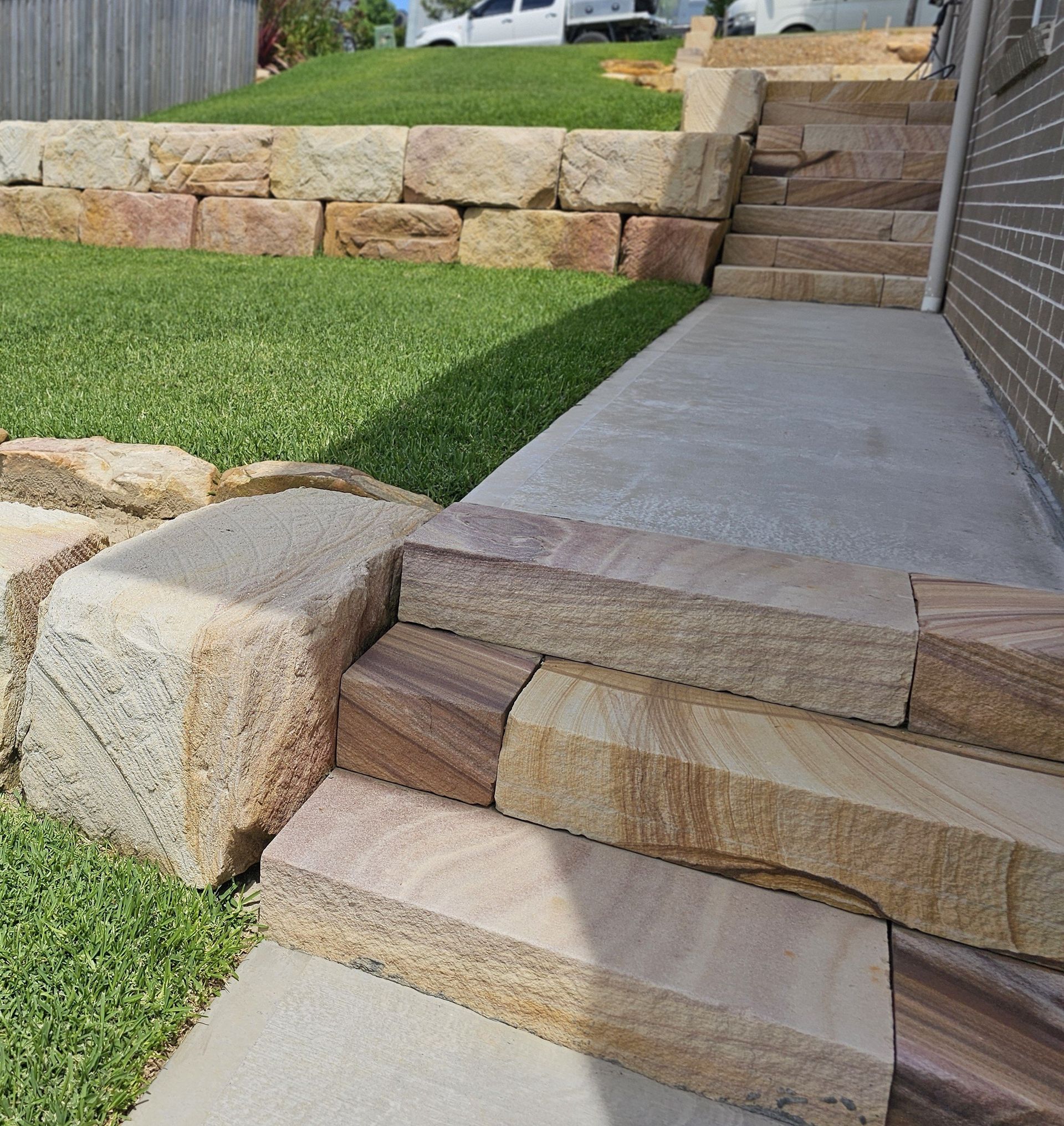 Stone retaining wall and steps leading to a concrete path and doorway, bordered by green grass.