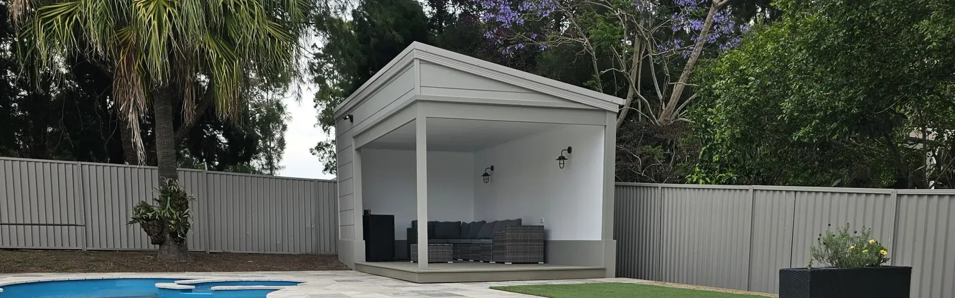 White-walled poolside cabana with seating area, nestled between trees and a white picket fence.