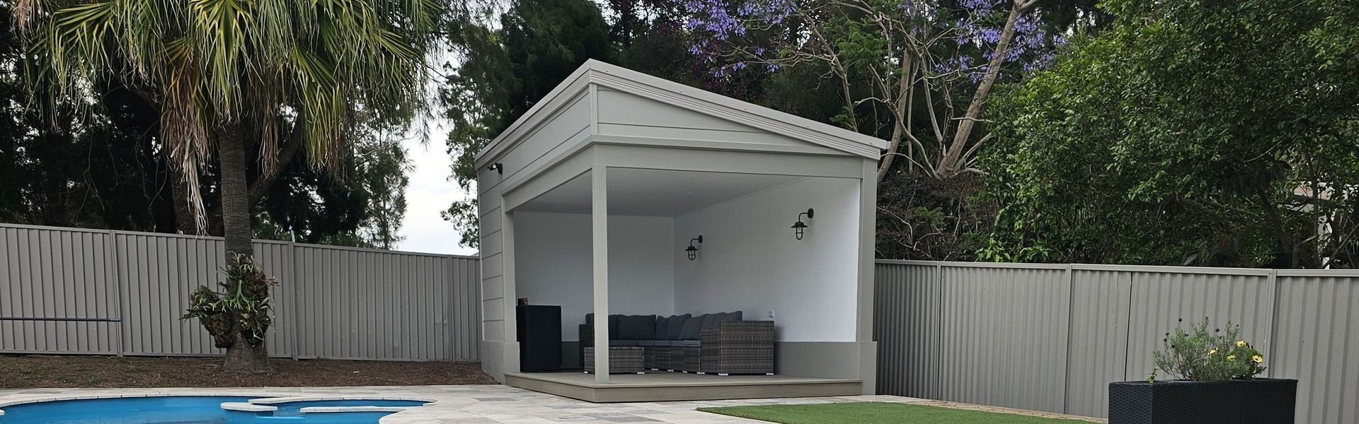 White gazebo beside a pool and lawn, with a fence and trees in the background.