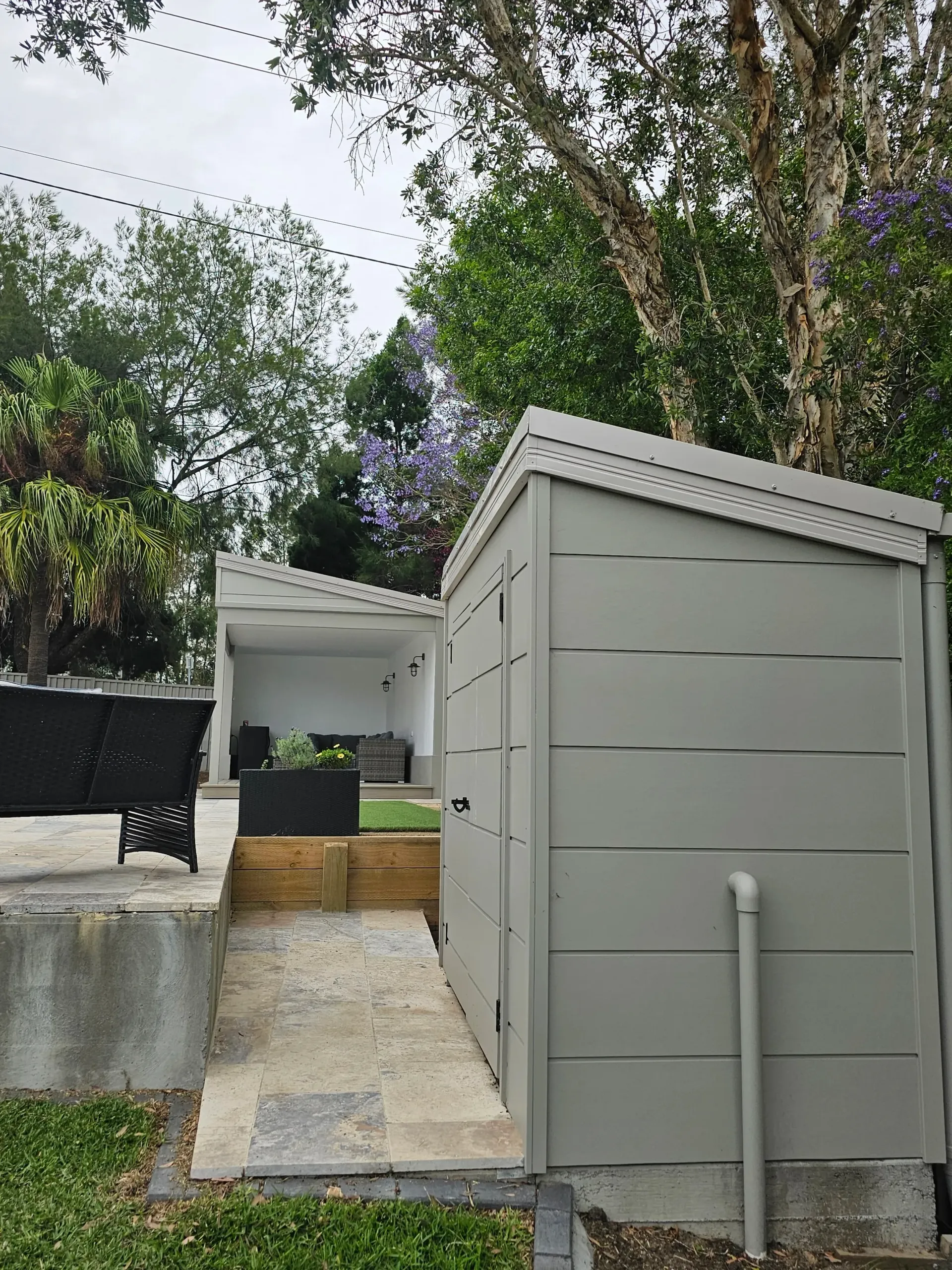 Gray shed with a slanted roof next to a patio area with a white structure and lush greenery.
