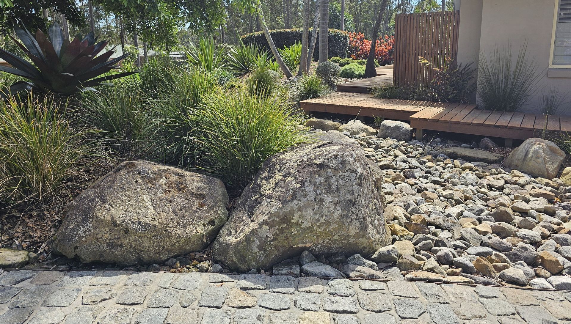 Large rocks and gravel in a landscaped garden, with plants, a wooden deck, and a building in the background.
