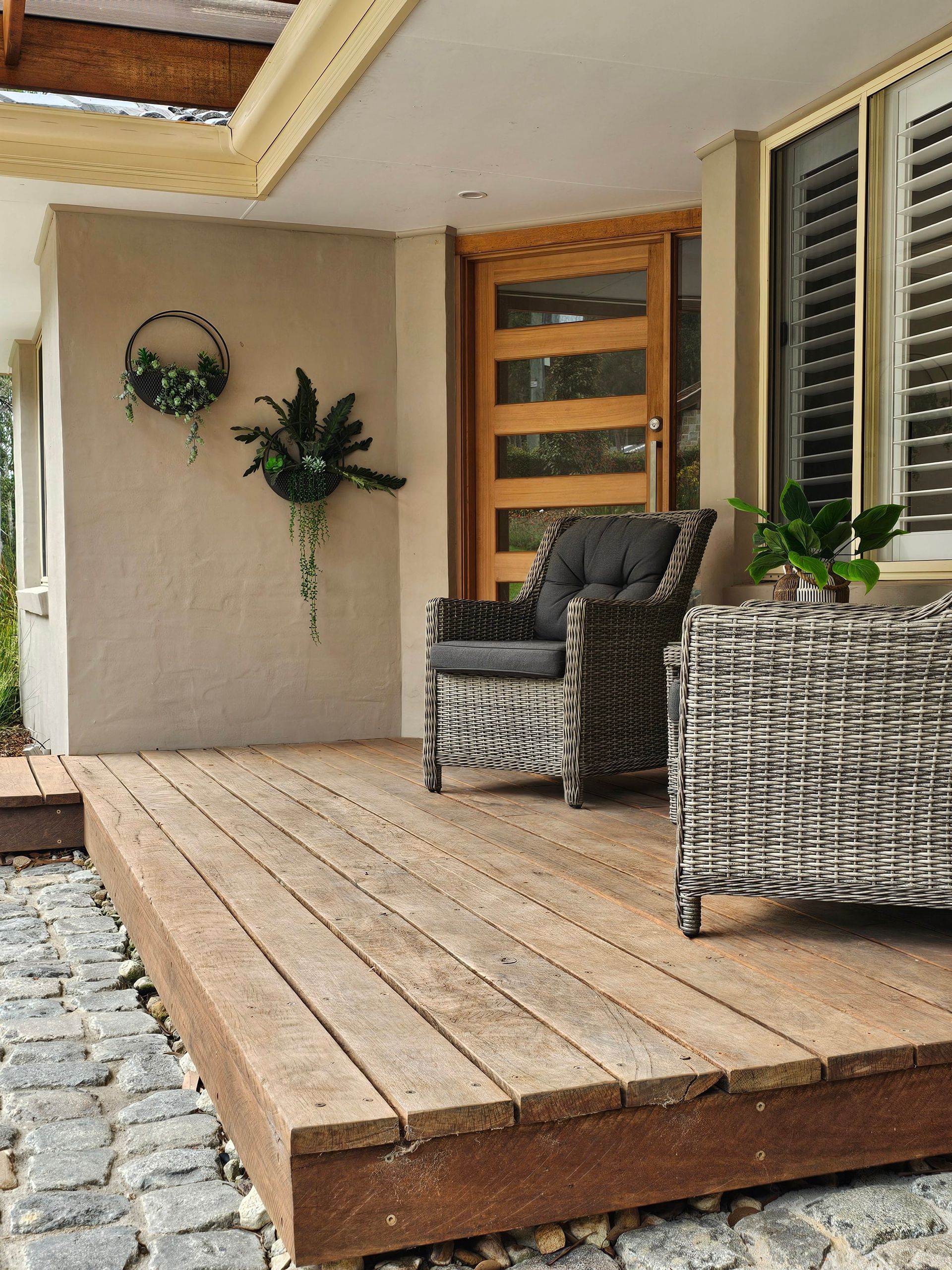 Wooden deck with wicker chairs, a doorway, and greenery. Beige walls, cobblestone edging.