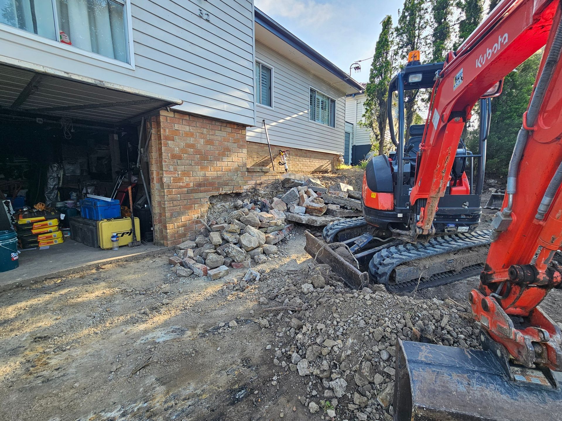 Orange excavator digging near a two-story house with exposed brick foundation.