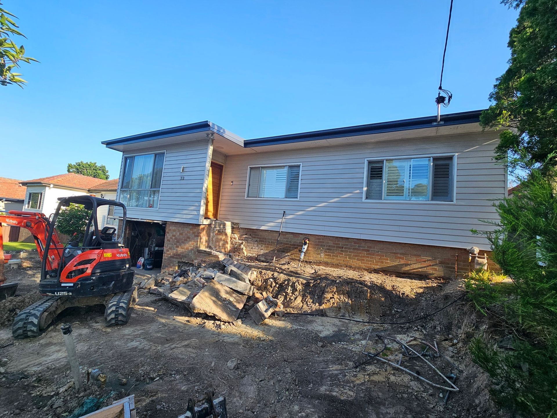 House on a hillside under construction with an excavator nearby; beige siding, blue sky.
