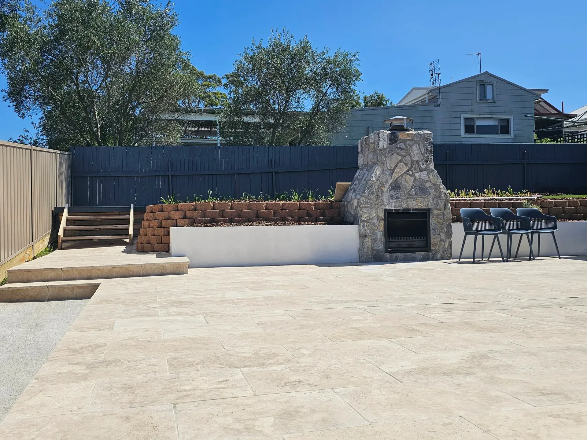 Backyard patio with fireplace, seating, and steps leading to a raised area, set against a fence and trees.