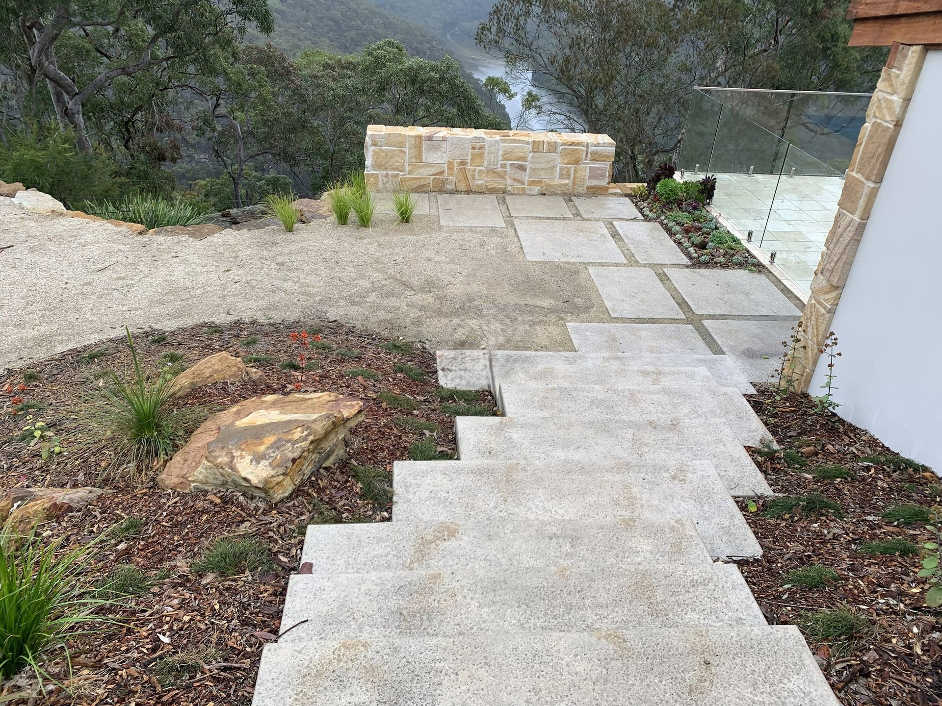 Stone steps and pathway leading up to a stone wall and glass railing, with gravel and landscaping on either side.