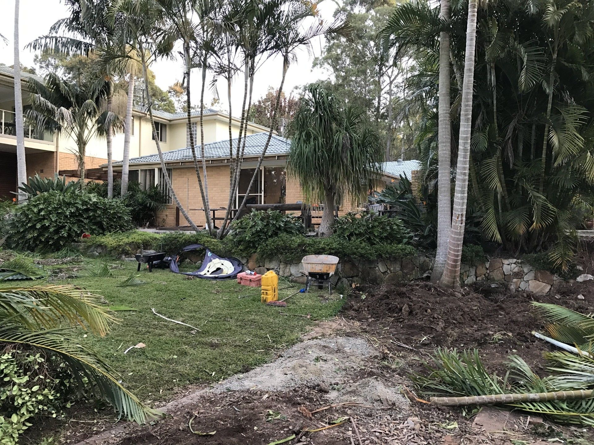 House with tan brick exterior, surrounded by trees and bushes. Tools and debris in yard.