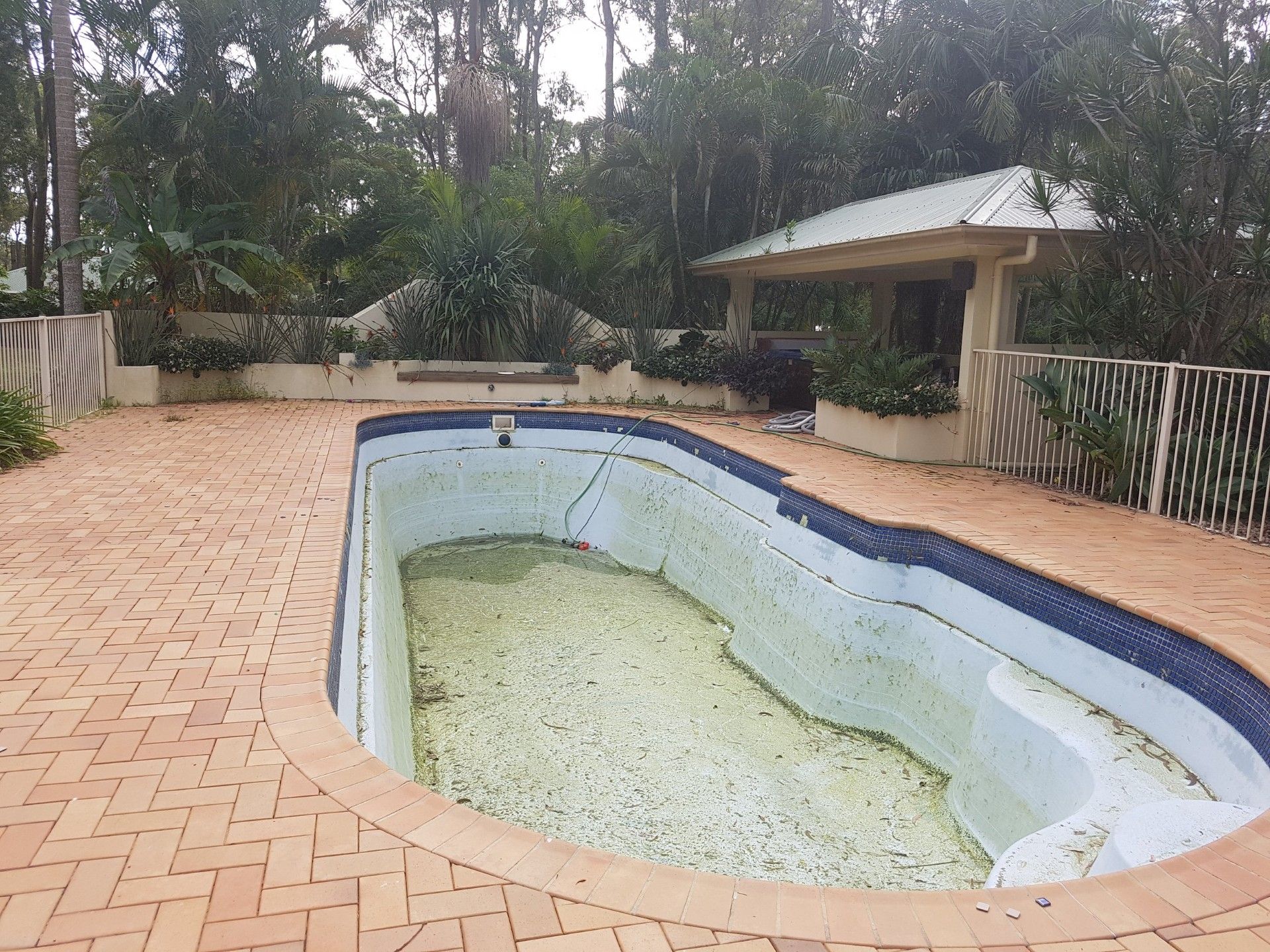 Empty, algae-covered swimming pool surrounded by brick patio, trees, and small cabana.