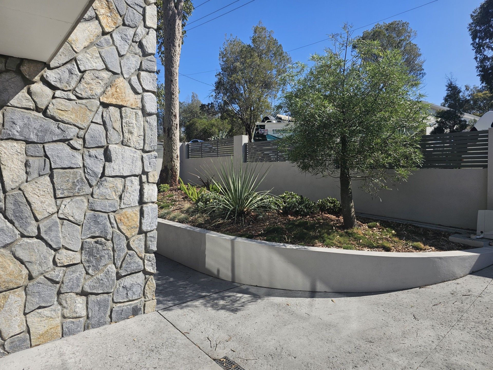 Stone wall beside concrete driveway, landscaped garden bed with small trees and shrubs, sunny day.
