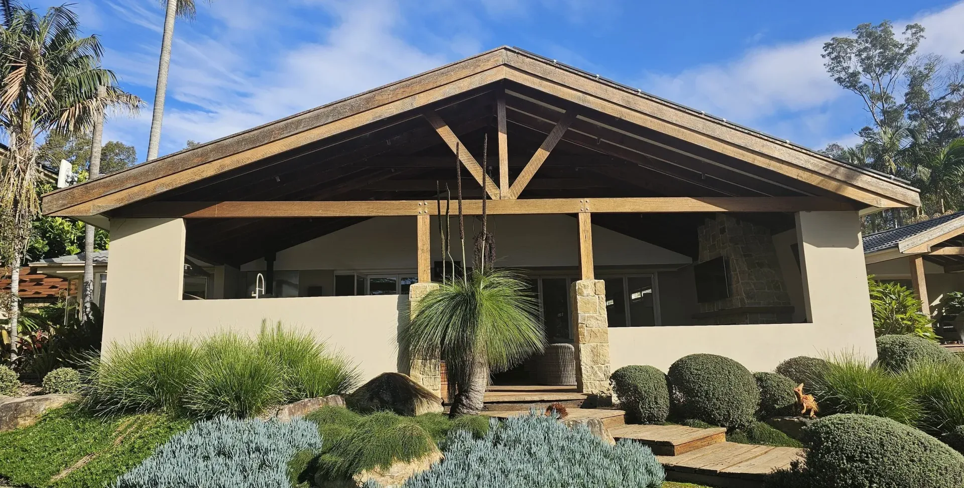 Tan stucco house with wooden porch and roof, surrounded by landscaping, under blue sky.
