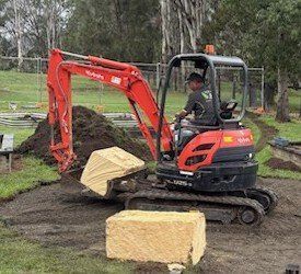 Orange excavator moving a large rectangular block. A person is operating the machine outdoors.