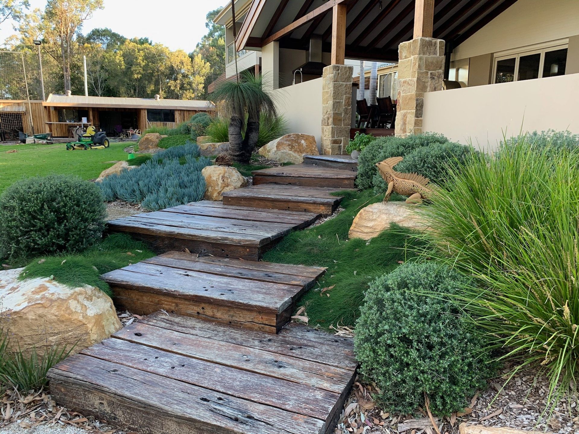 Wooden steps ascend a landscaped garden leading to a building's entrance. Green plants and rocks surround the steps.